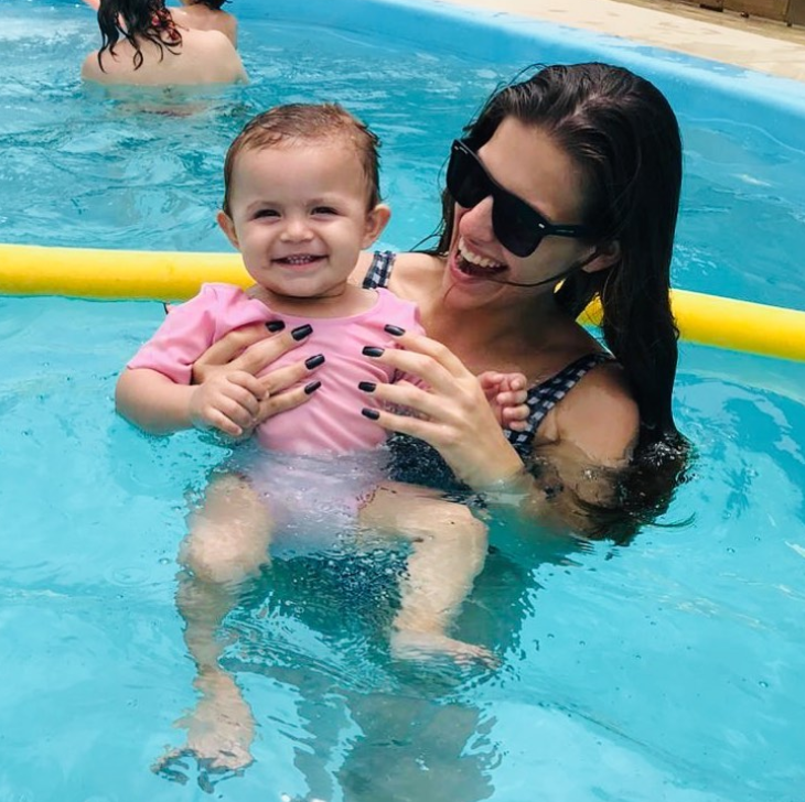 Mãe e filha brincando na piscina. A menina está no colo da mãe sorrindo enquanto a mãe olha sorrindo para o bebê. 